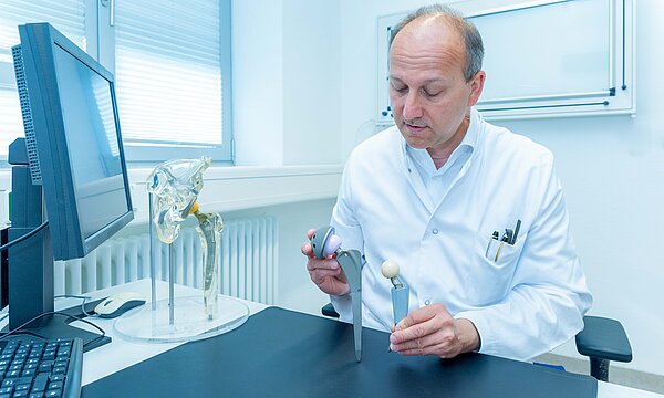 Ein Chefarzt des Städtischen Klinikums Karlsruhe sitzt an einem Tisch und hat verschiedene Gelenkprothesen in der Hand.