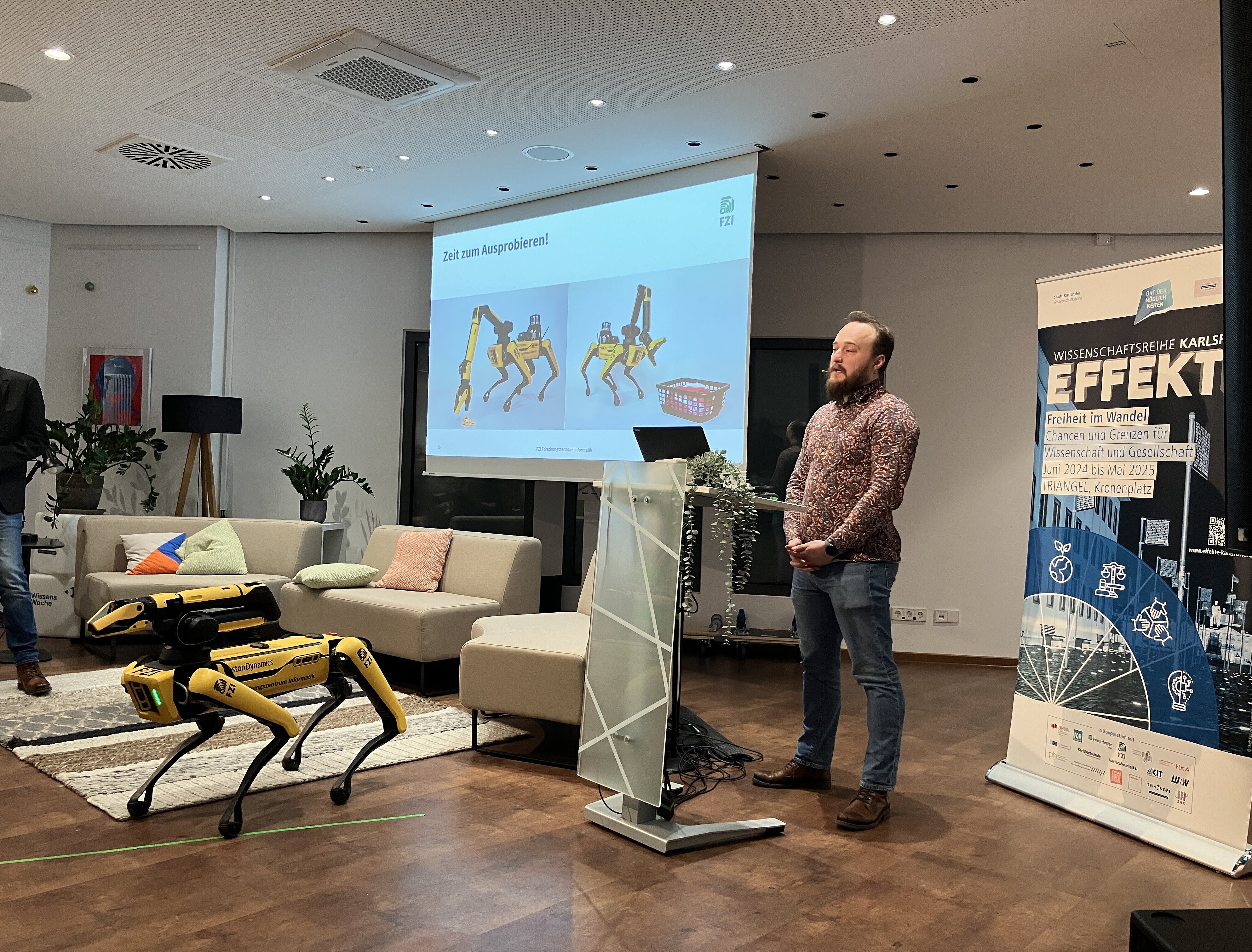 Ein Forscher des FZI steht vor einem Rednerpult im Rahmen der Veranstaltung EFFEKTE. Neben ihm steht ein gelber Roboter, der die Form eines Hundes hat und Spot genannt wird.