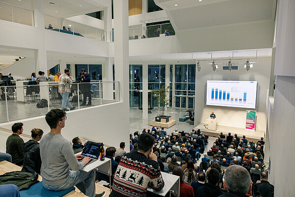 Viele Menschen sitzen in einem großen Saal und schauen auf eine Leinwand, auf der ein Vortrag projiziert wird.