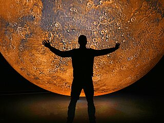Ein Mensch steht vor einem riesigen rötlichen Planeten (Mars vermutlich)