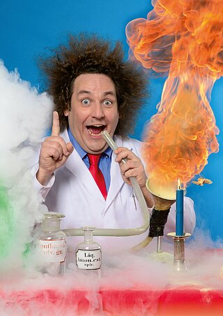 Ein Wissenschaftler mit Einsteinfrisur hinter einem Tisch auf dem Dampf und Feuer herrschen. Die Experimente darstellen sollen.